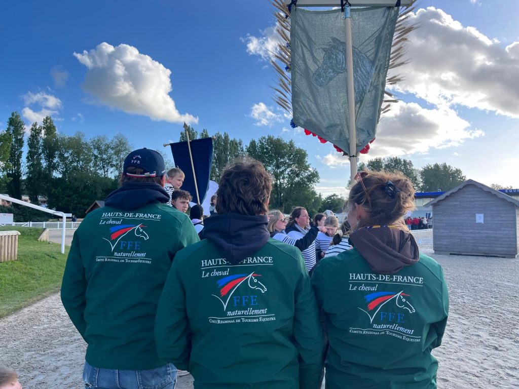 3 personnes de dos avec des vestes vertes floquées du logo officiel des CRTE "Le cheval, naturellement". Une des personnes porte une bannière représentant une tête de cheval avec des brins de blés et et fleurs sur le pourtour.