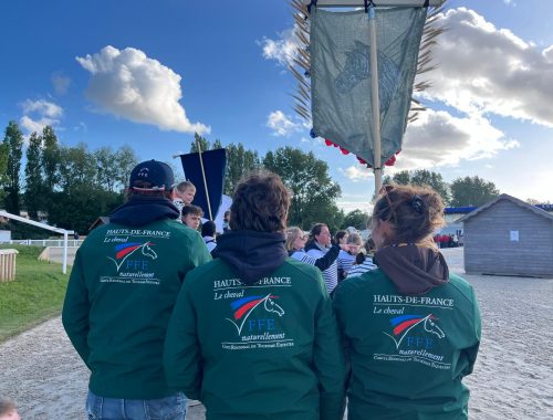 3 personnes de dos avec des vestes vertes floquées du logo officiel des CRTE "Le cheval, naturellement". Une des personnes porte une bannière représentant une tête de cheval avec des brins de blés et et fleurs sur le pourtour.